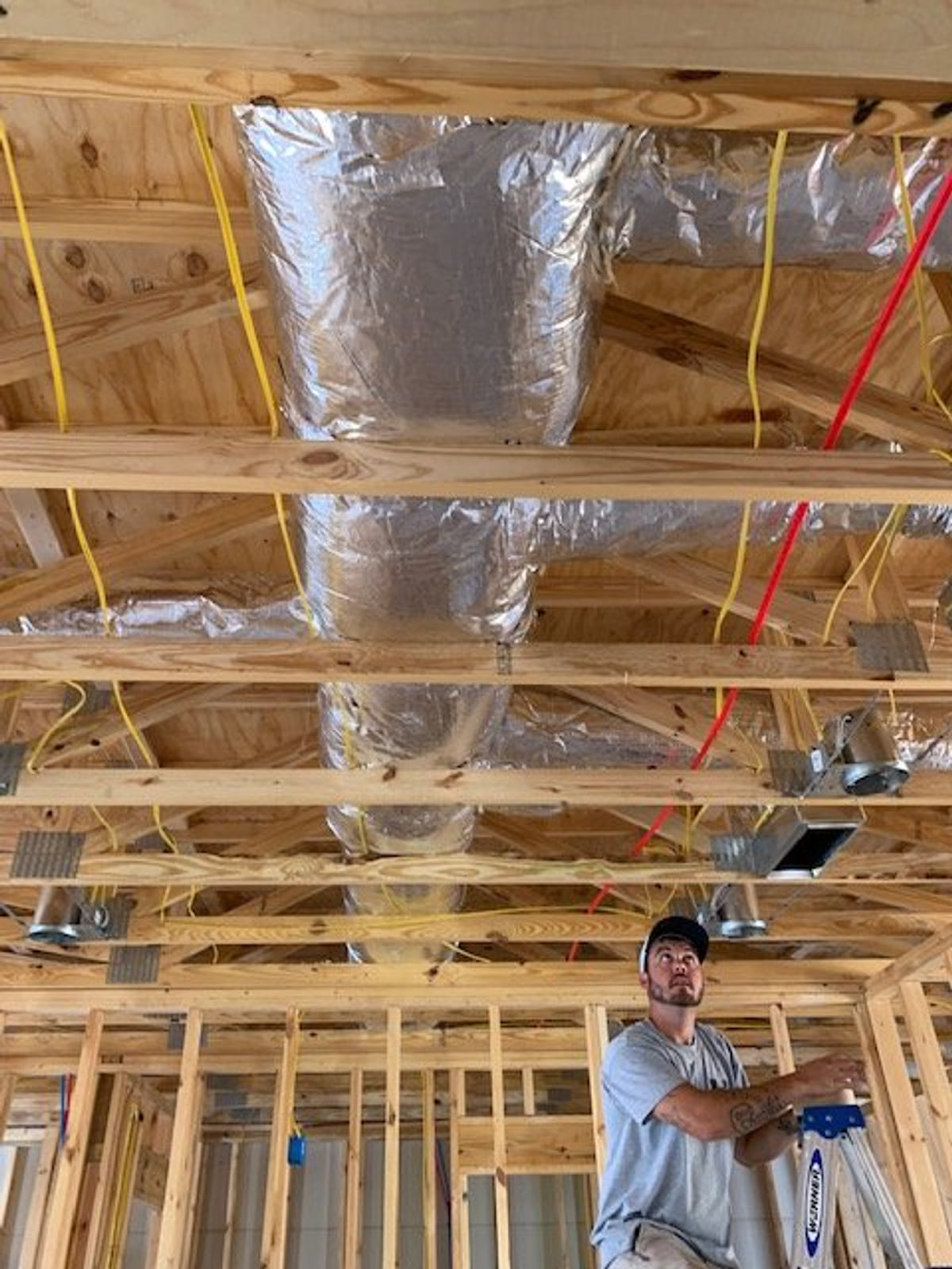 HVAC technician working beneath foil-faced ductwork in a wood-framed building.